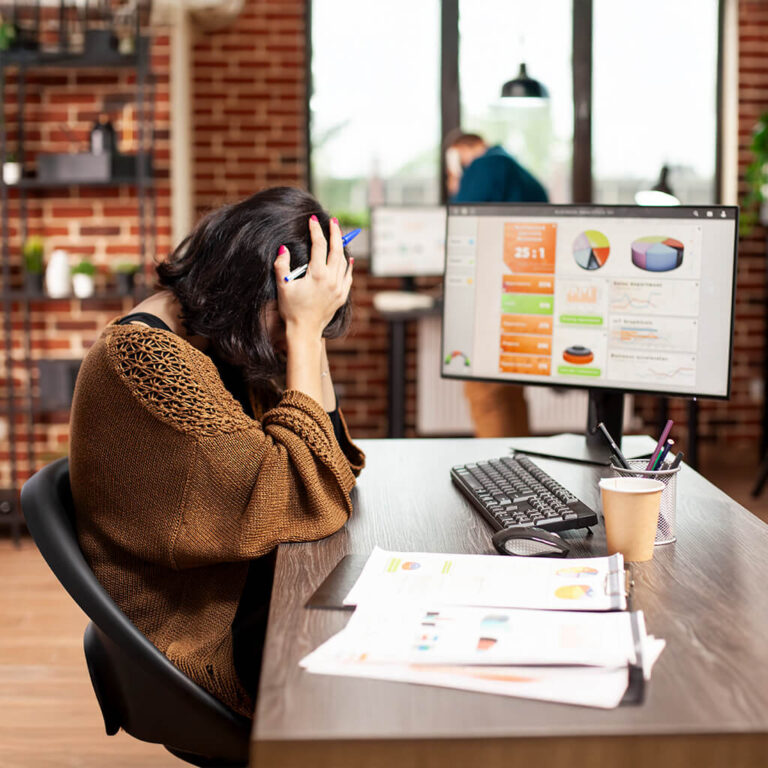 An overwhelmed marketer or business owner rests their head in their hands at a desk, with performance data visible on the monitor behind them.