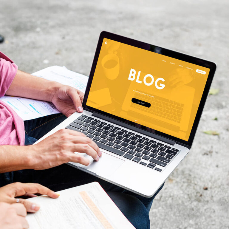 Close-up of hands typing on a laptop that displays a website page with the word "BLOG" prominently. This image illustrates the concept of writing content that drives leads.
